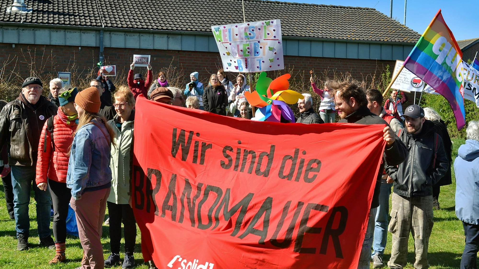 Das Bild zeigt Demonstrierende. Zwei von ihnen halten ein Banner mit der Aufschrift „Wir sind die Brandmauer“.