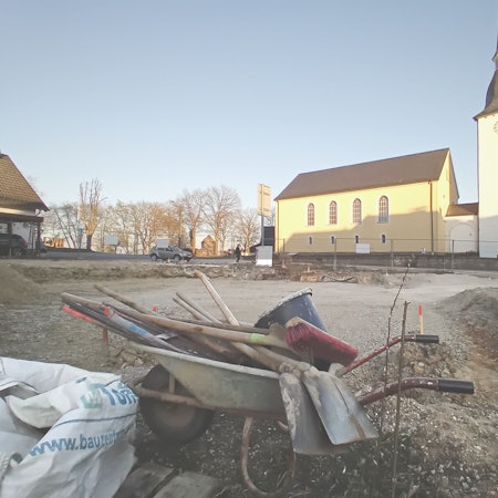 Vorn eine Schubkarre mit Werkzeug, hinten eine Kirche.