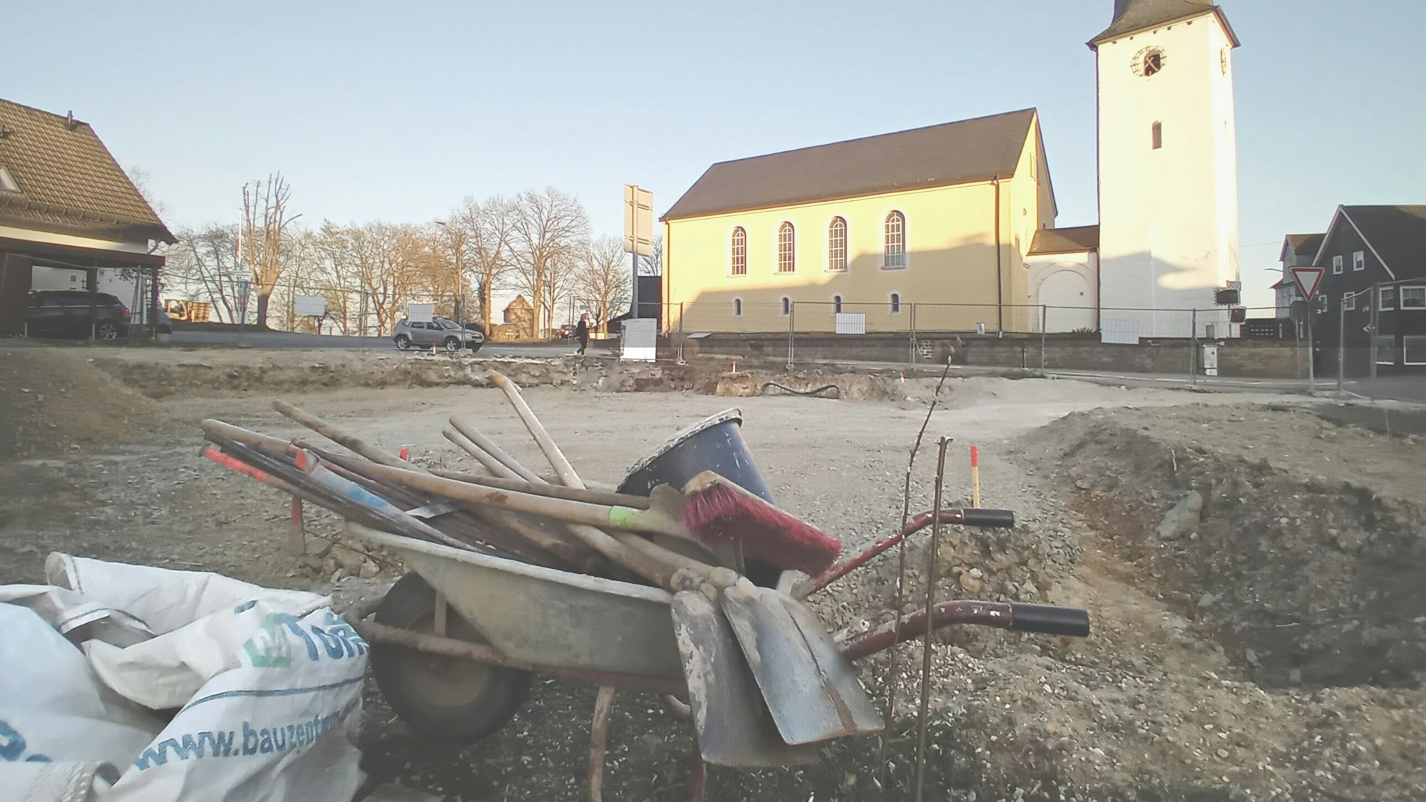 Vorn eine Schubkarre mit Werkzeug, hinten eine Kirche.