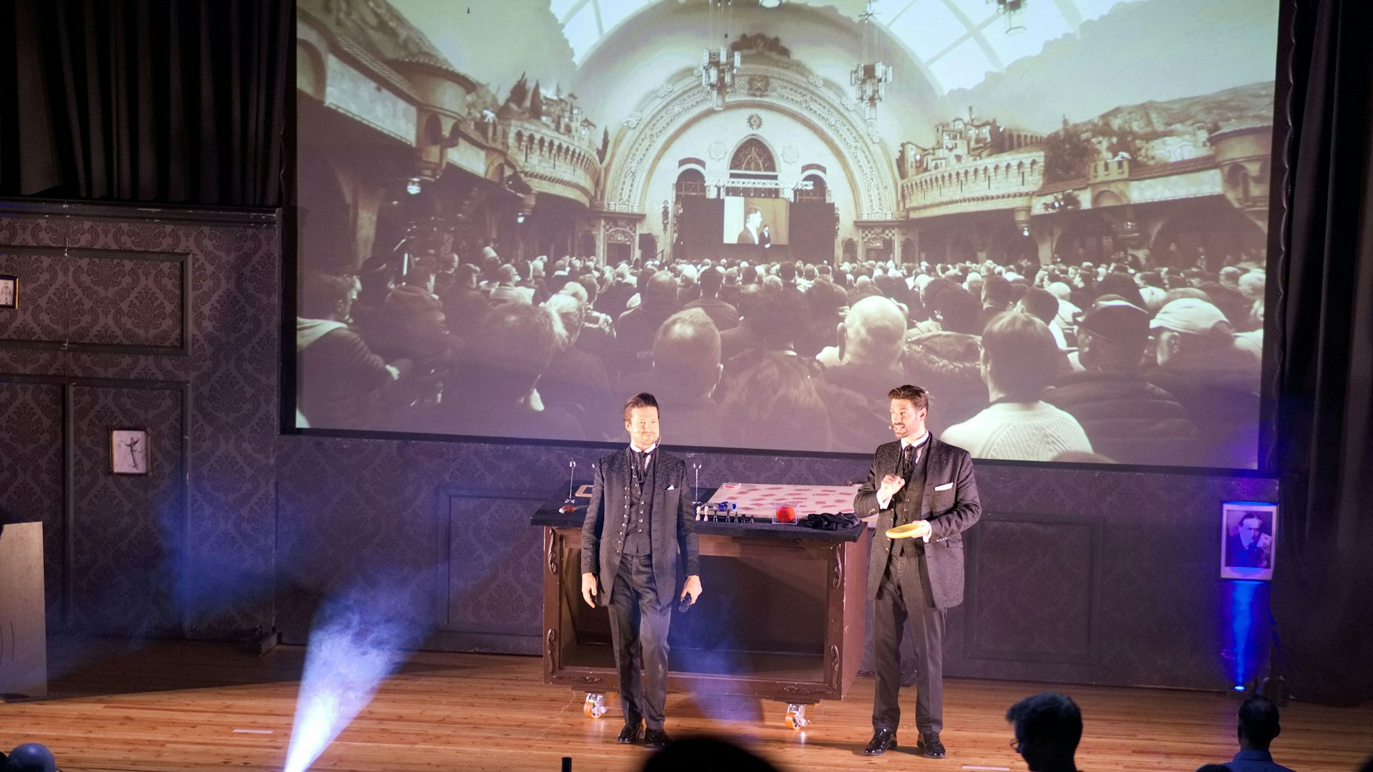 Das Bild zeigt die beiden Männer auf der Bühne. Sie stehen vor einer großen Leinwand mit einem Schwarz-Weiß-Foto, auf dem ein voll besetzter Theatersaal zu sehen ist.