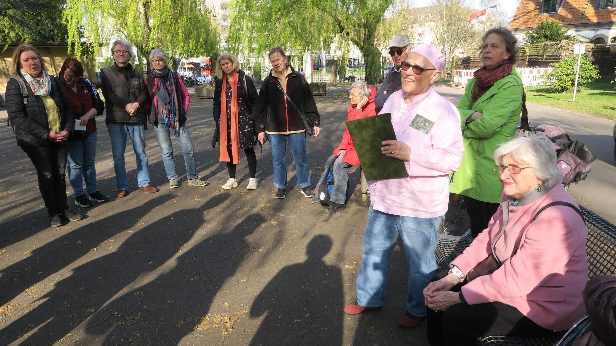 Menschen stehen im Halbkreis auf einem großen Vorplatz, eine Frau mit rosa Mütze referiert.