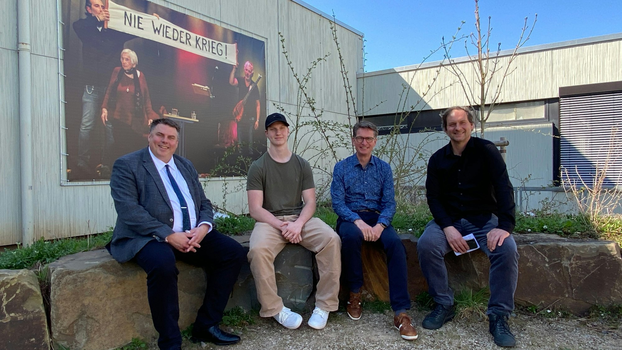 Im Erinnerungsgarten der Marienschule sitzen Michael Mombaur (v.l.), Leander Dehner, Denis Rolf und Florian Bethe. Im Hintergrund ist ein Plakat befestigt mit der Aufschrift „Nie wieder Krieg!“.