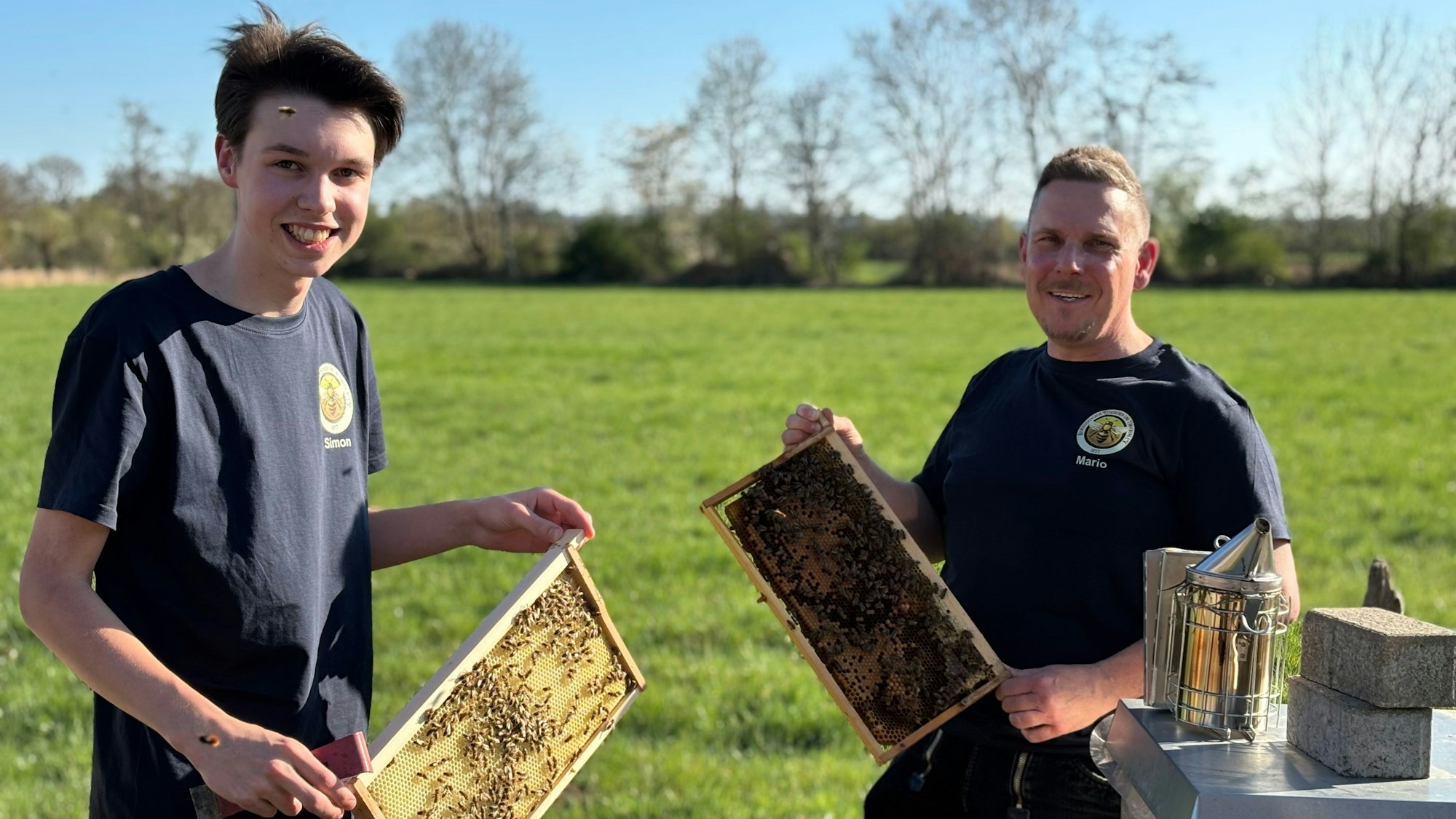 Die beiden Imker halten Bienenwaben in die Luft. Der jüngere Mann wird von Bienen umschwirrt. Sie stehen vor einer Wiese mit Bäumen.