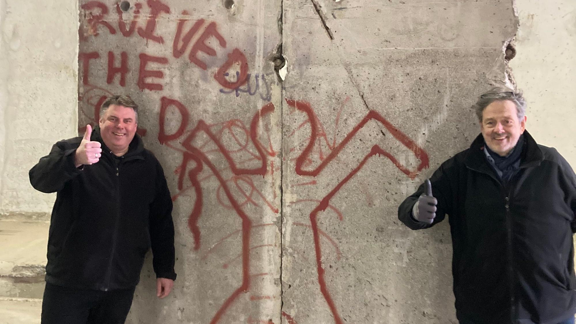 Zwei Männer in schwarzen Jacken stehen an einem Segment der Berliner Mauer. Sie jecken jeweils einen Daumen nach oben.
