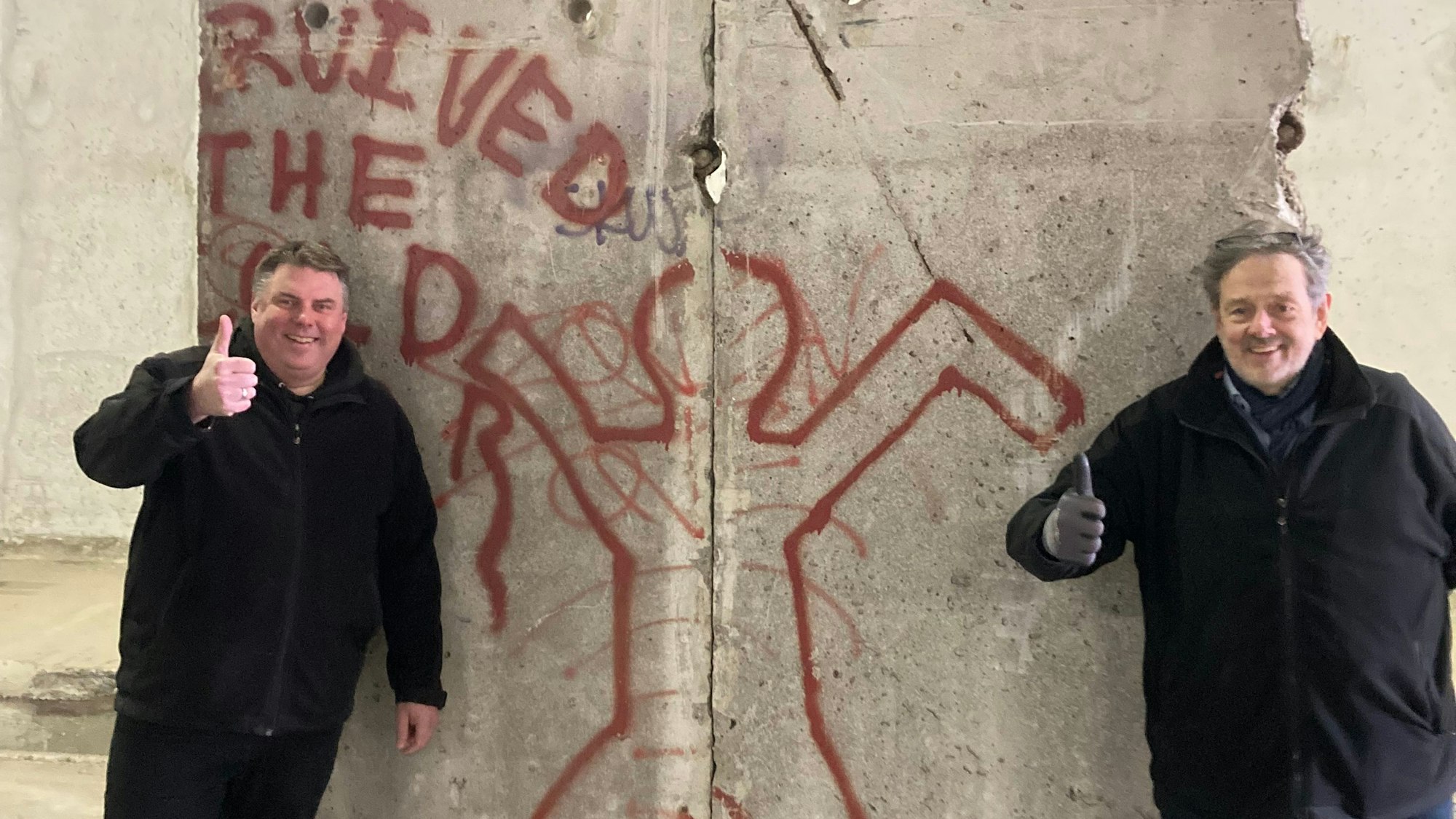 Zwei Männer in schwarzen Jacken stehen an einem Segment der Berliner Mauer. Sie jecken jeweils einen Daumen nach oben.