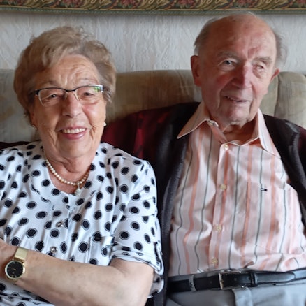 Ein älteres Ehepaar sitzt nebeneinander auf einem Sofa und lächelt.