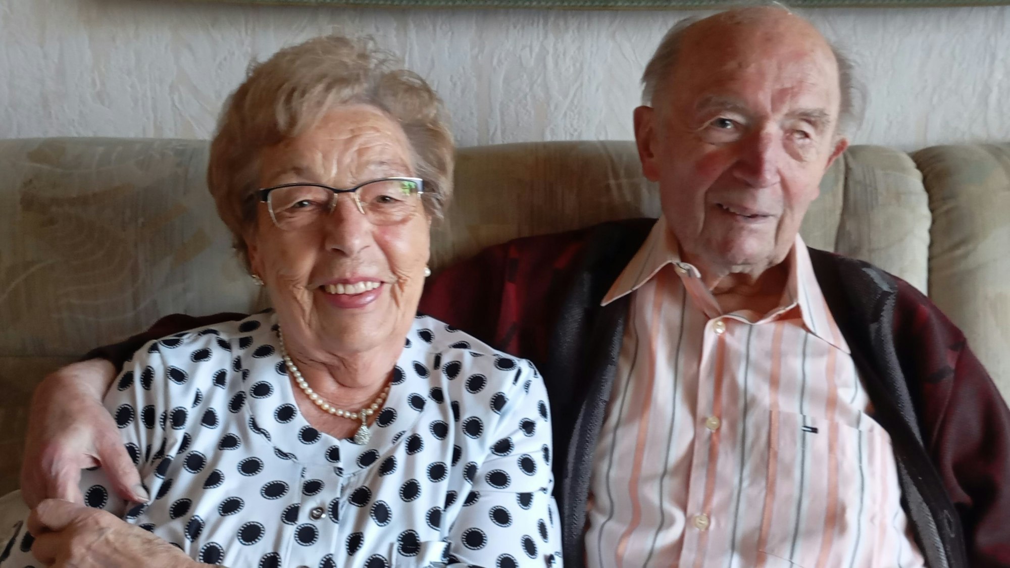 Ein älteres Ehepaar sitzt nebeneinander auf einem Sofa und lächelt.