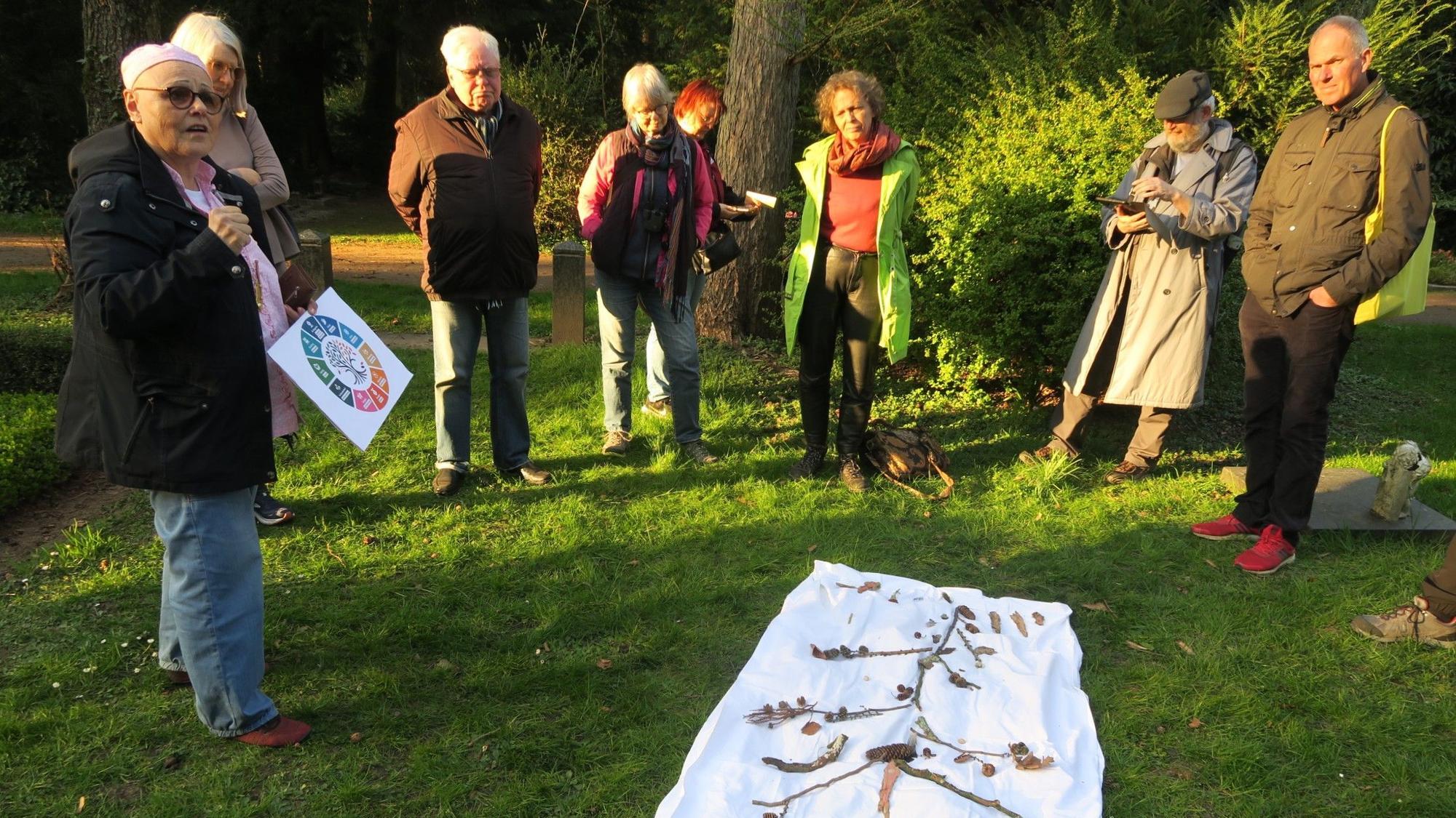 Menschen stehen im Halbkreis auf einem Stück Rasen, auf einem Tuch haben sie Zweige und Blätter zu einem Baum angeordnet.