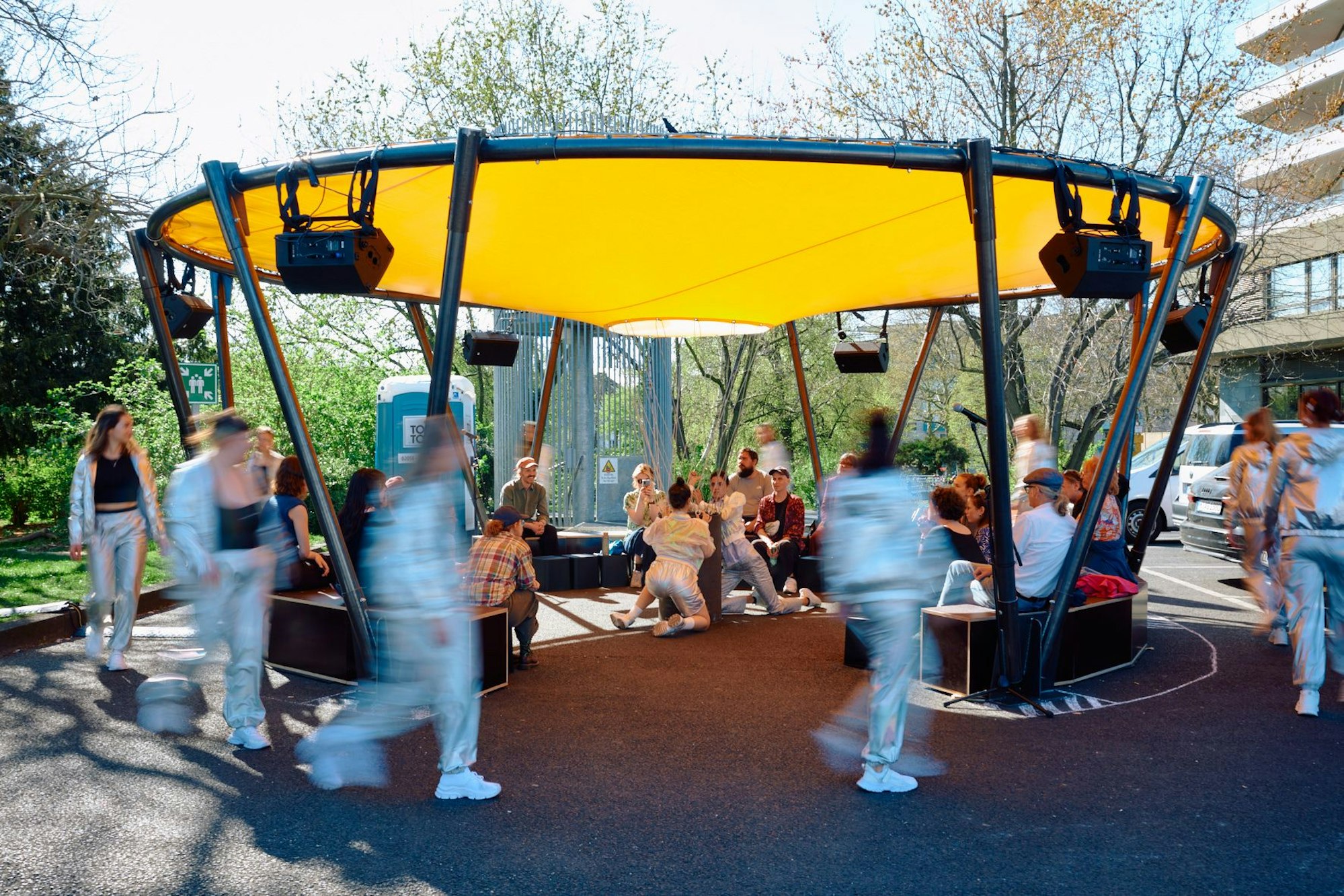Schauspieler in silbernen Anzügen laufen im Kreis um die Stahl-Installation herum.