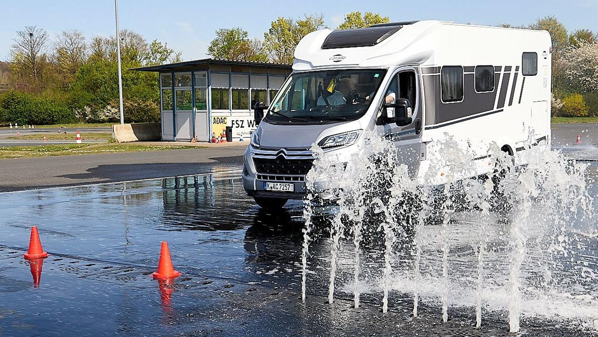 Ein Campingwagen fährt durch eine große Pfütze. Wasser spritzt aus dem Boden.