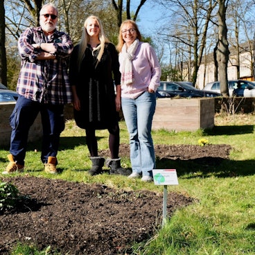 Zwei Frauen und ein Mann stehen vor einem Beet, in dem nur eine winzige Pflanze zu sehen ist.