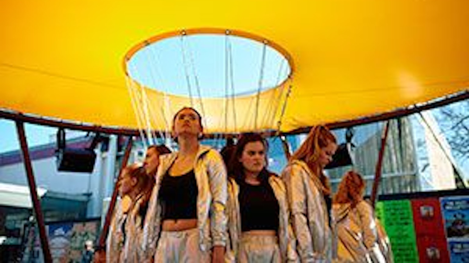 Mädchen in silbernen Trainingsanzügen und schwarzen Tops stehen unter einem gelben Baldachin, der an einer Stahlkonstruktion angebracht ist.