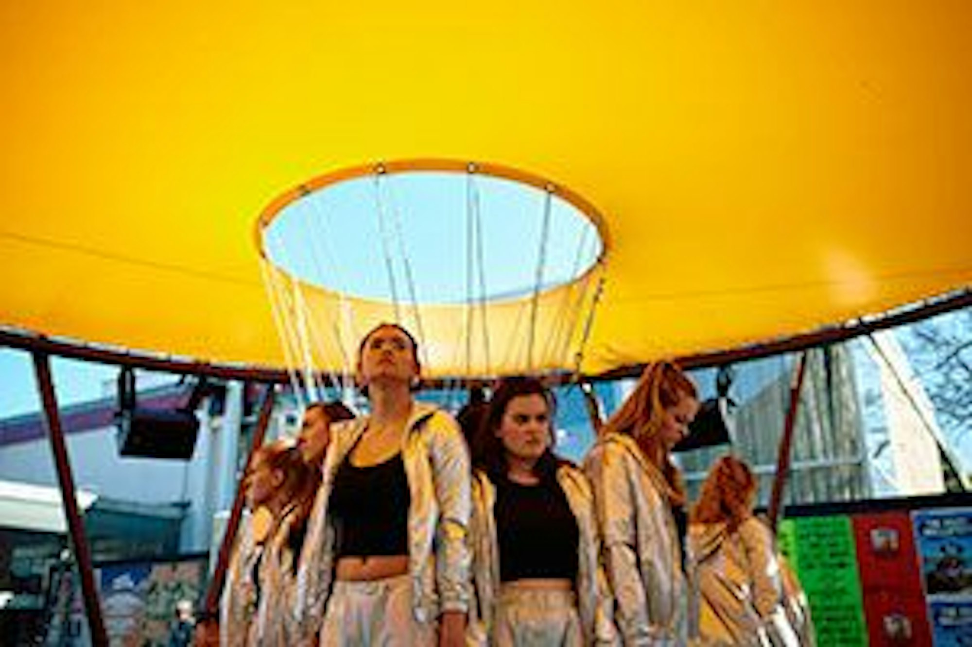 Mädchen in silbernen Trainingsanzügen und schwarzen Tops stehen unter einem gelben Baldachin, der an einer Stahlkonstruktion angebracht ist.