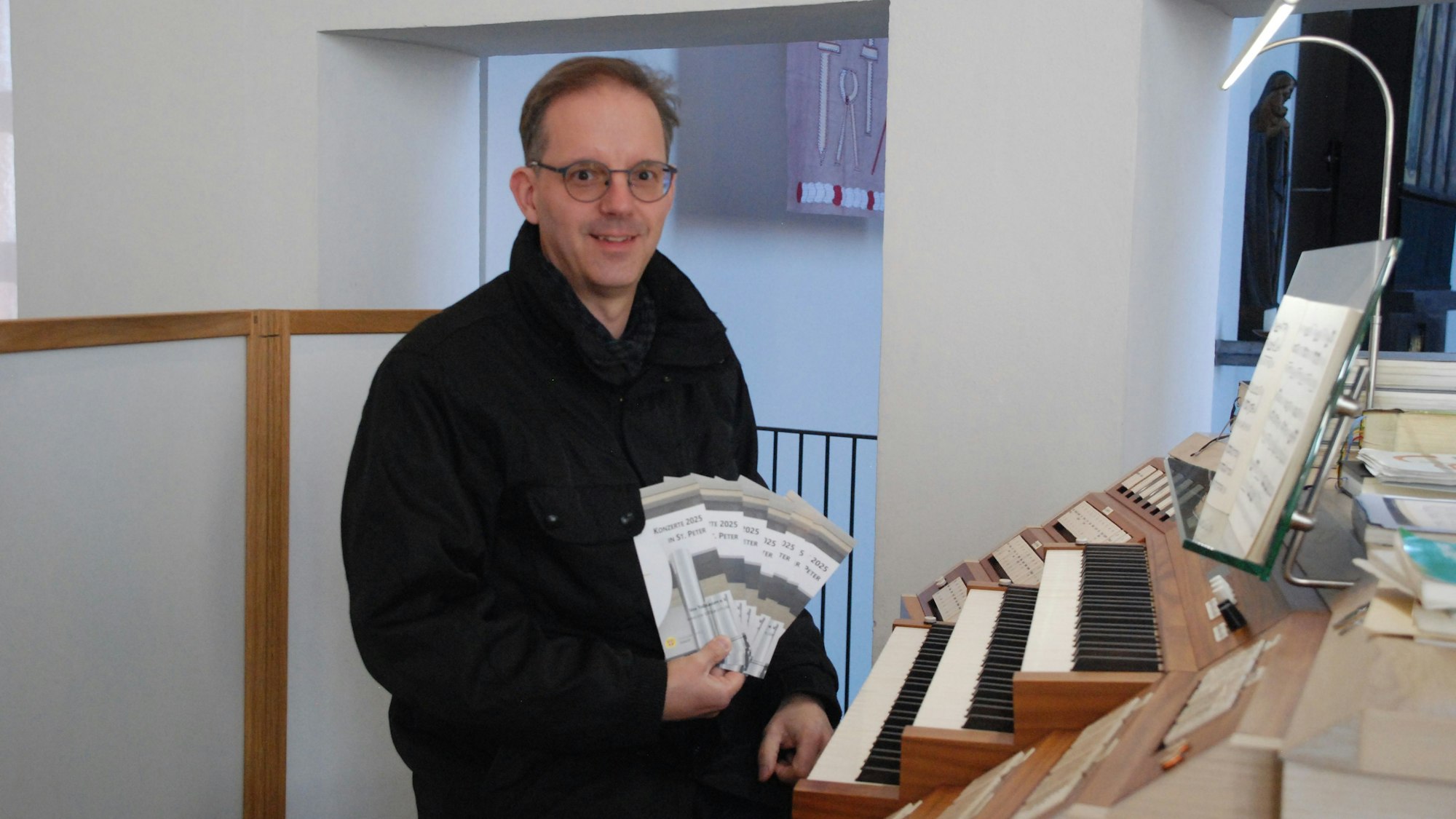 Ein Mann, der Brille und eine schwarze Jacke trägt, hält aufgefächerte Veranstaltungshefte in seiner rechten Hand.