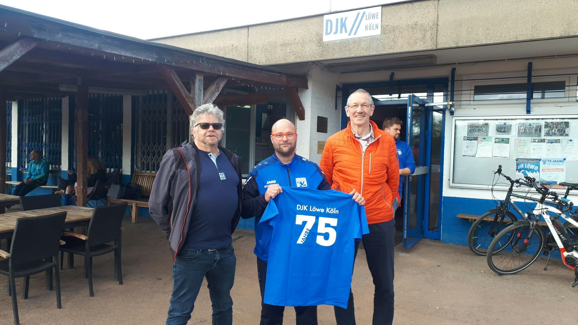 Drei Männer stehen nebeneinander, der in der Mitte hält ein Trikot, auf dem „DJK Löwe Köln, 75 Jahre“ steht.