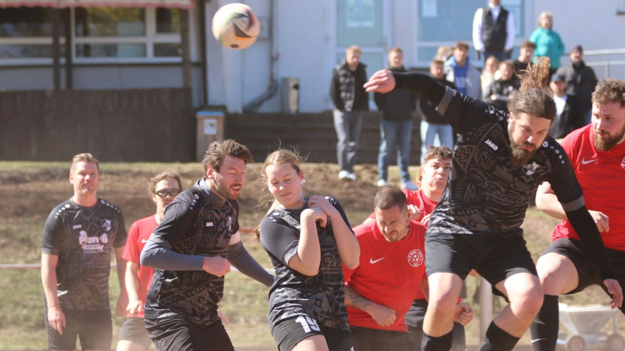 Eine Spielerin und mehrere Spieler beider Mannschaft springen, überwiegend mit geschlossenen Augen, zum Kopfball hoch.