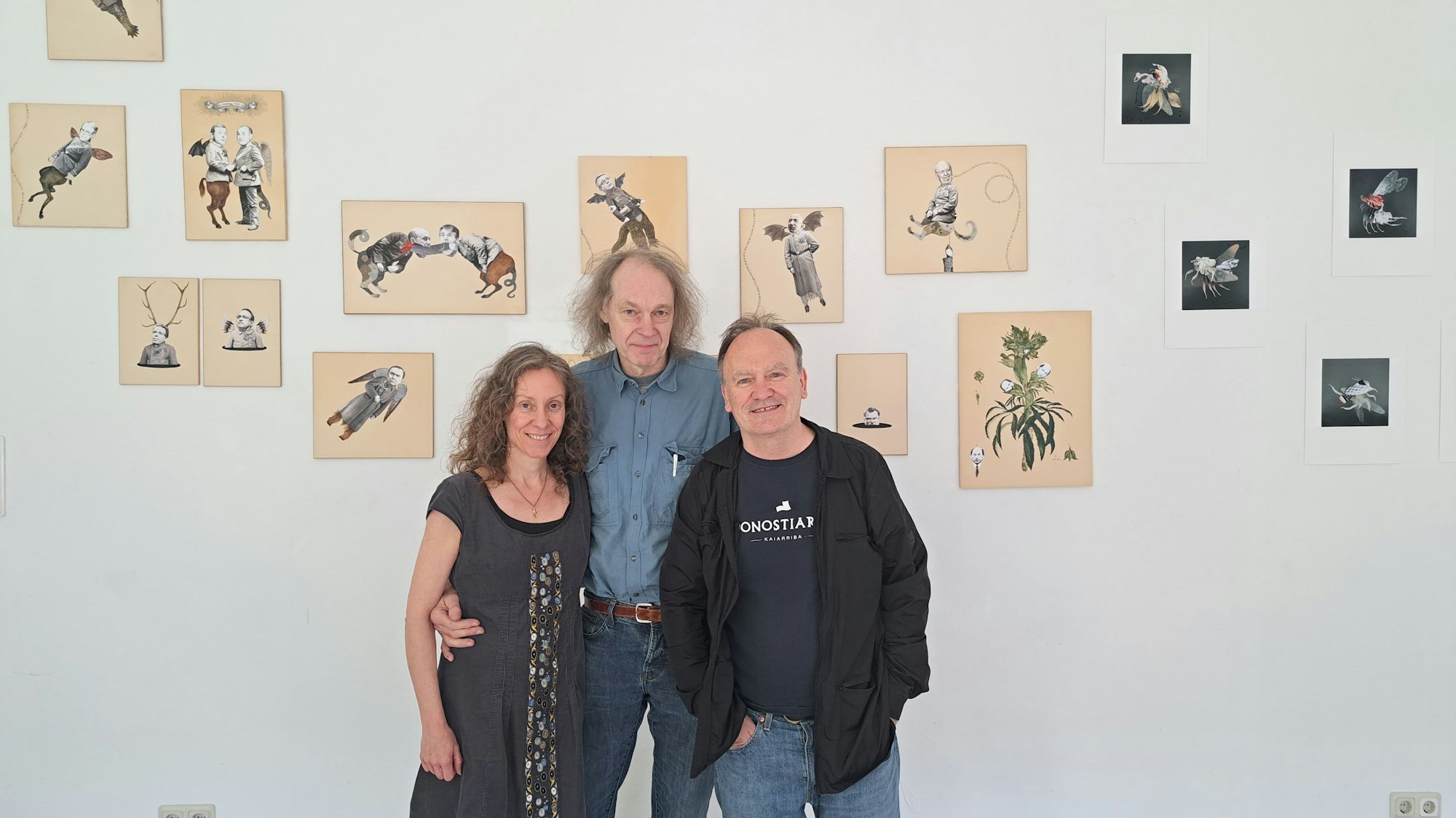 Eine Frau und zwei Männer stehen nebeneinander vor einer Wand, an der Bilder hängen.