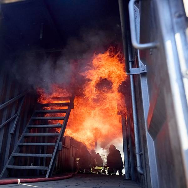 Drei Feuerwehrleute hocken in einer Brandsimulationsanlage auf dem Boden. Über ihnen sind Flammen.