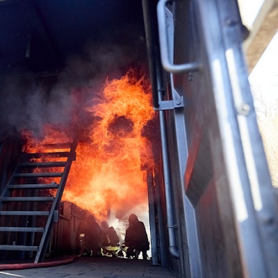 Drei Feuerwehrleute hocken in einer Brandsimulationsanlage auf dem Boden. Über ihnen sind Flammen.
