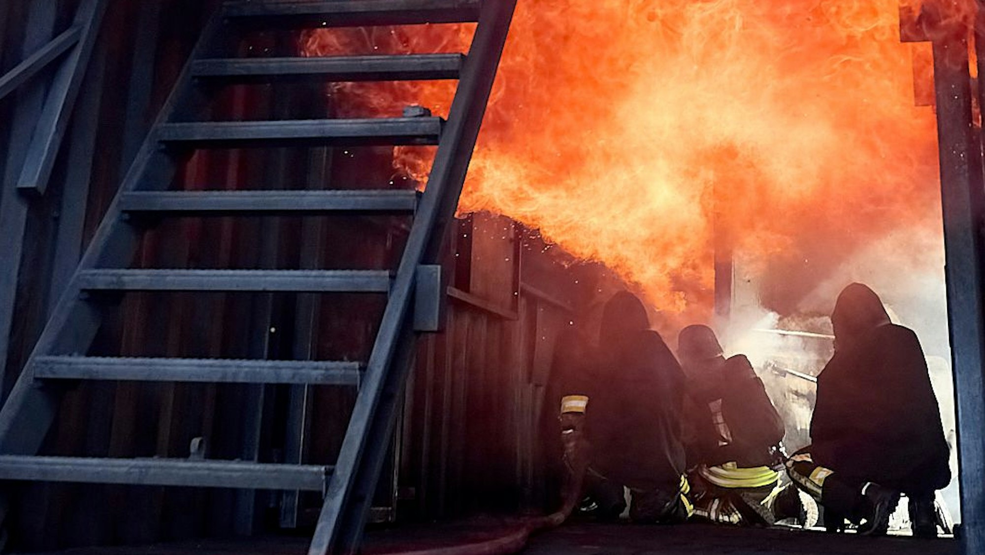 Drei Feuerwehrleute hocken in einer Brandsimulationsanlage auf dem Boden. Über ihnen sind Flammen.