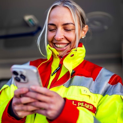 Das Bild zeigt Lea Pilger in ihrer Rettungsdienstkleidung. Sie hält ein Smartphone in der Hand hat.