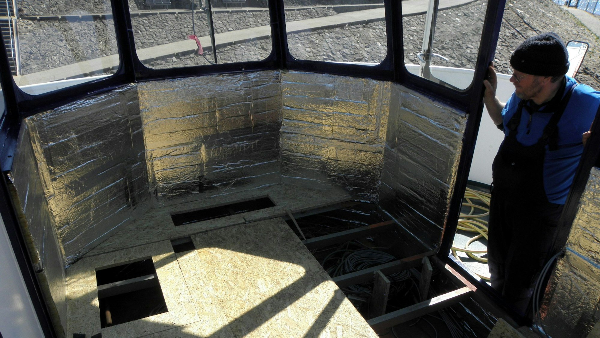 Ein Mann schaut in einen Raum mit vielen Fenstern, von dem aus das Schiff auf dem Rursee gesteuert wird. Die Isolierung an den Wänden ist angebracht, die Einrichtung fehlt noch.