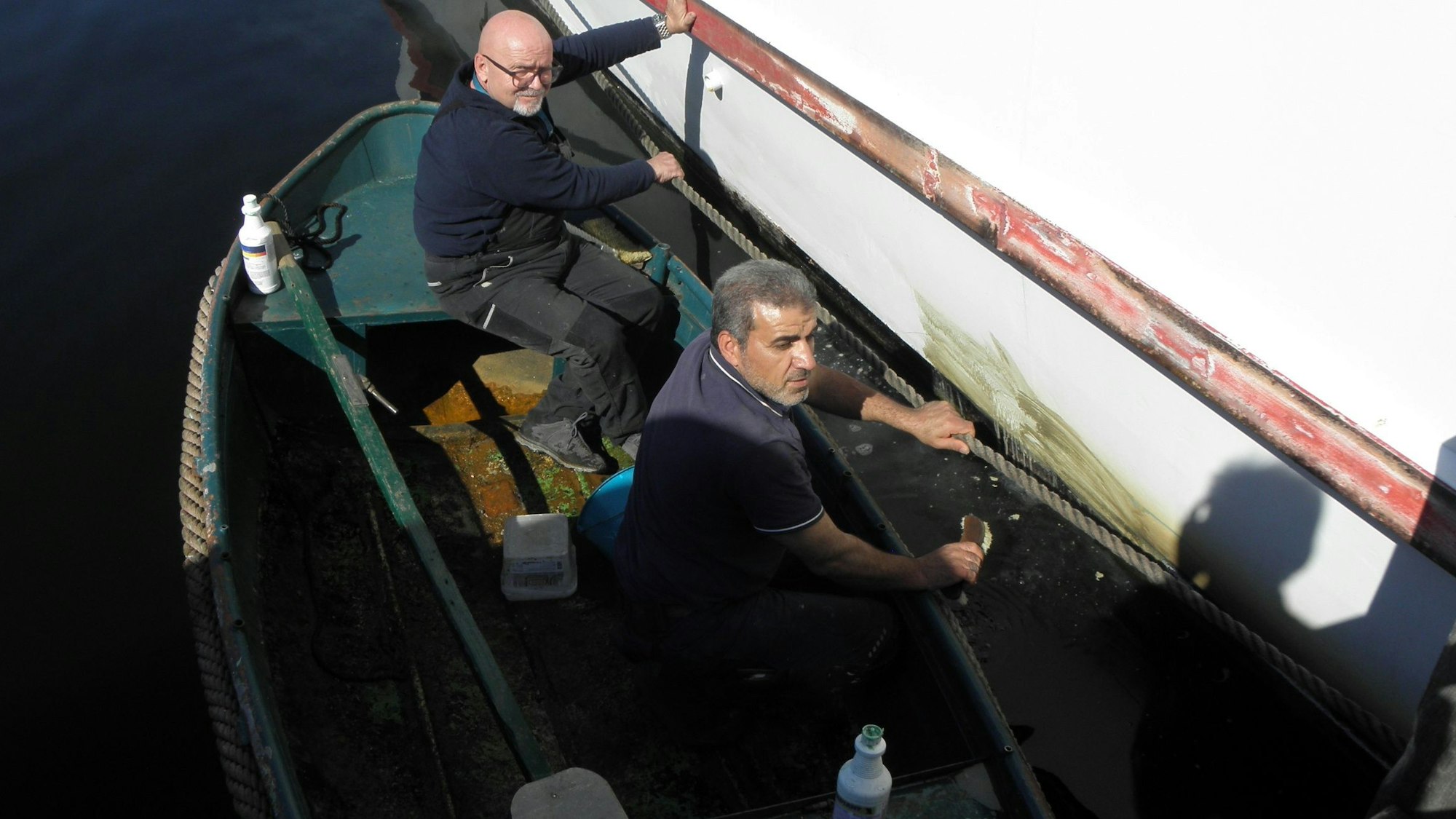 Zwei Männer sitzen in einem kleinen Ruderboot. Sie schrubben den grünen Algenbelag ab, der sich auf einem großen, weißen Schill gebildet hat.