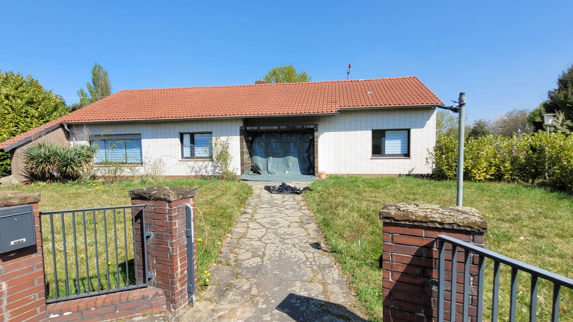 Ein in die Jahre gekommenes Haus mit heller Fassade, die Rolläden an den drei Fenstern sind heruntergelassen, dunkle Folie ersetzt die Eingangstür.