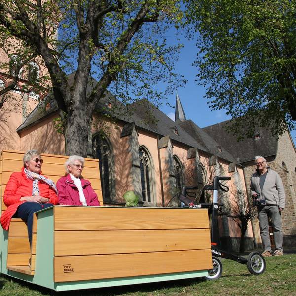 Zwei Frauen auf einem "Sofa", im Hintergrund die Pfarrkirche St. Johann Baptist.