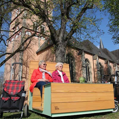 Zwei Frauen auf einem "Sofa", im Hintergrund die Pfarrkirche St. Johann Baptist.