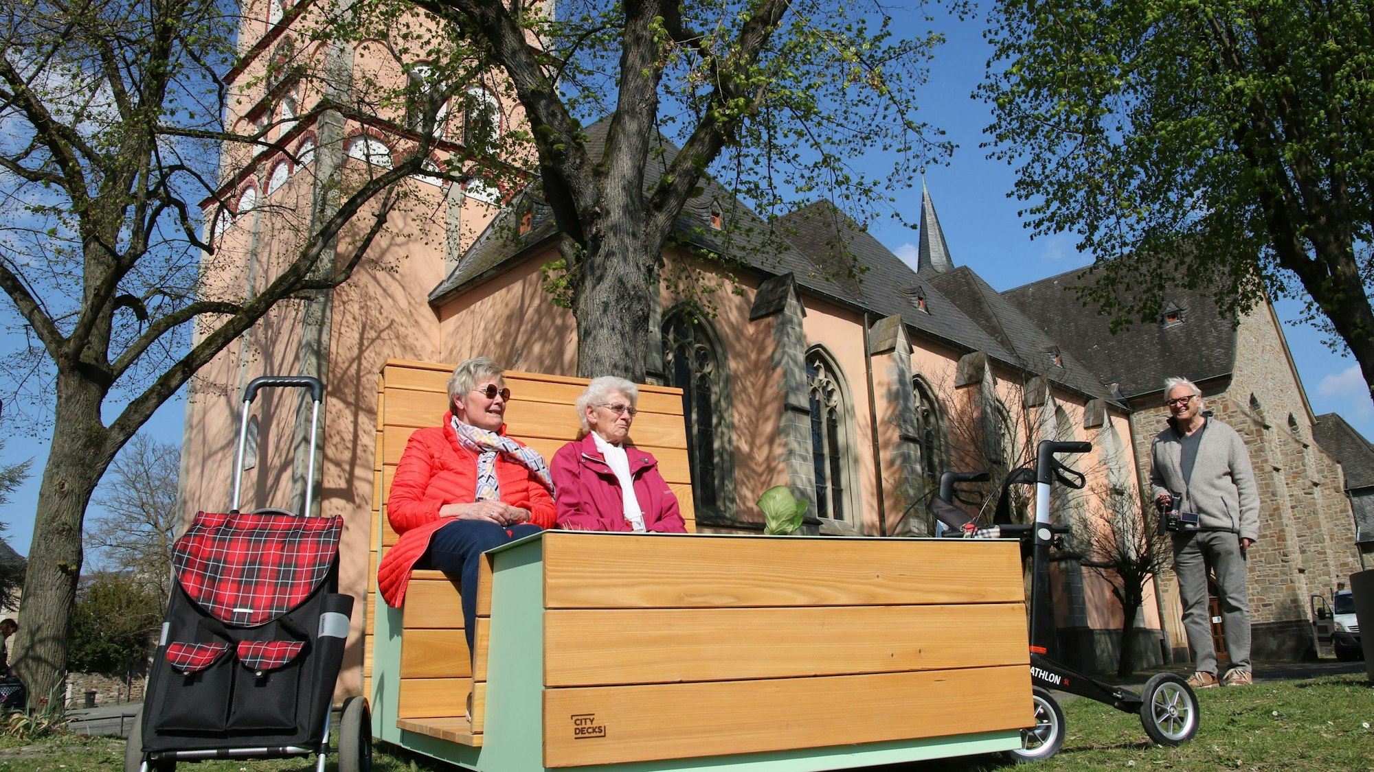 Zwei Frauen auf einem "Sofa", im Hintergrund die Pfarrkirche St. Johann Baptist.