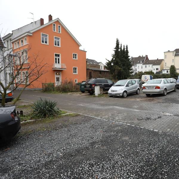 Parkende Autos auf einer Schotterfläche, links an der Seite ein Wohnhaus.