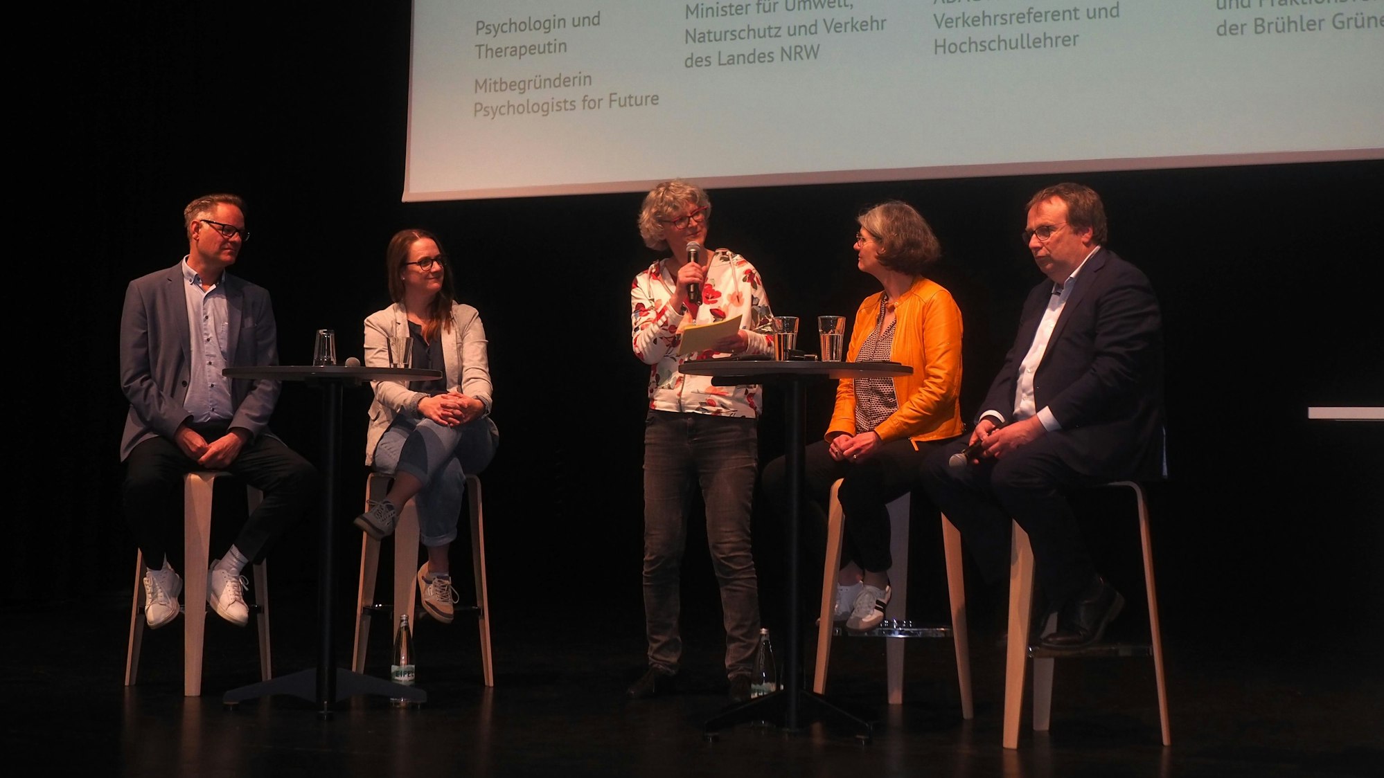 Männer und Frauen sitzen auf Hochstühlen auf einer Bühne.