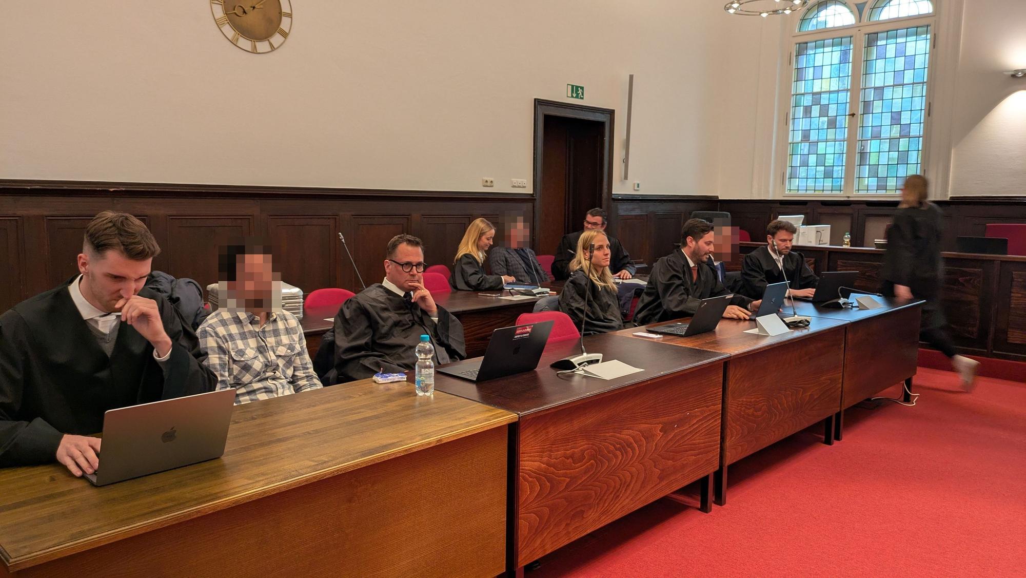 Die Angeklagten und Verteidiger sitzen in zwei Reihen an Tischen.