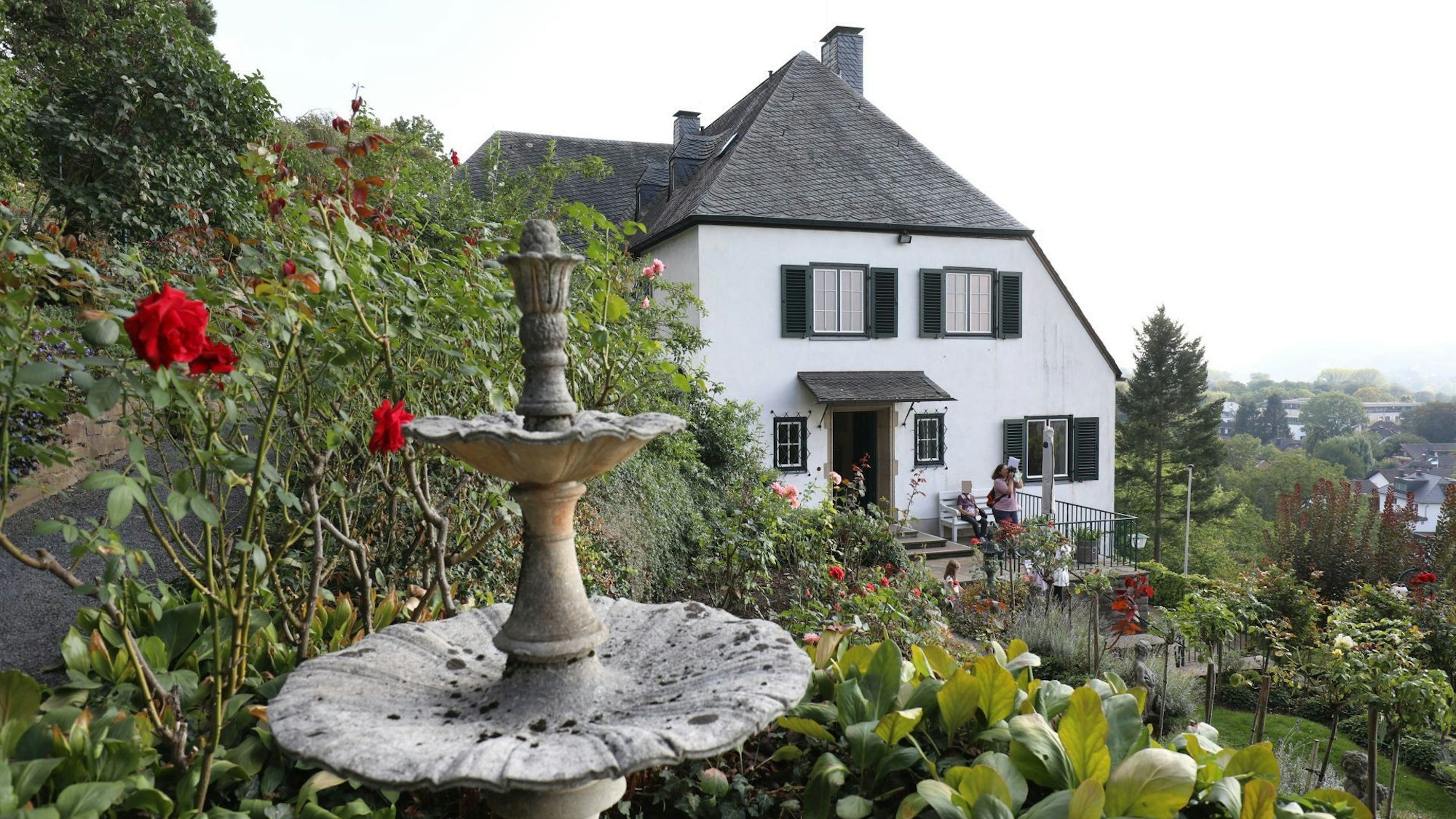 Ein Wohnhaus mit weißer Fassade und grünen Fensterläden, im Vordergrund Rosen in einem großen Garten.