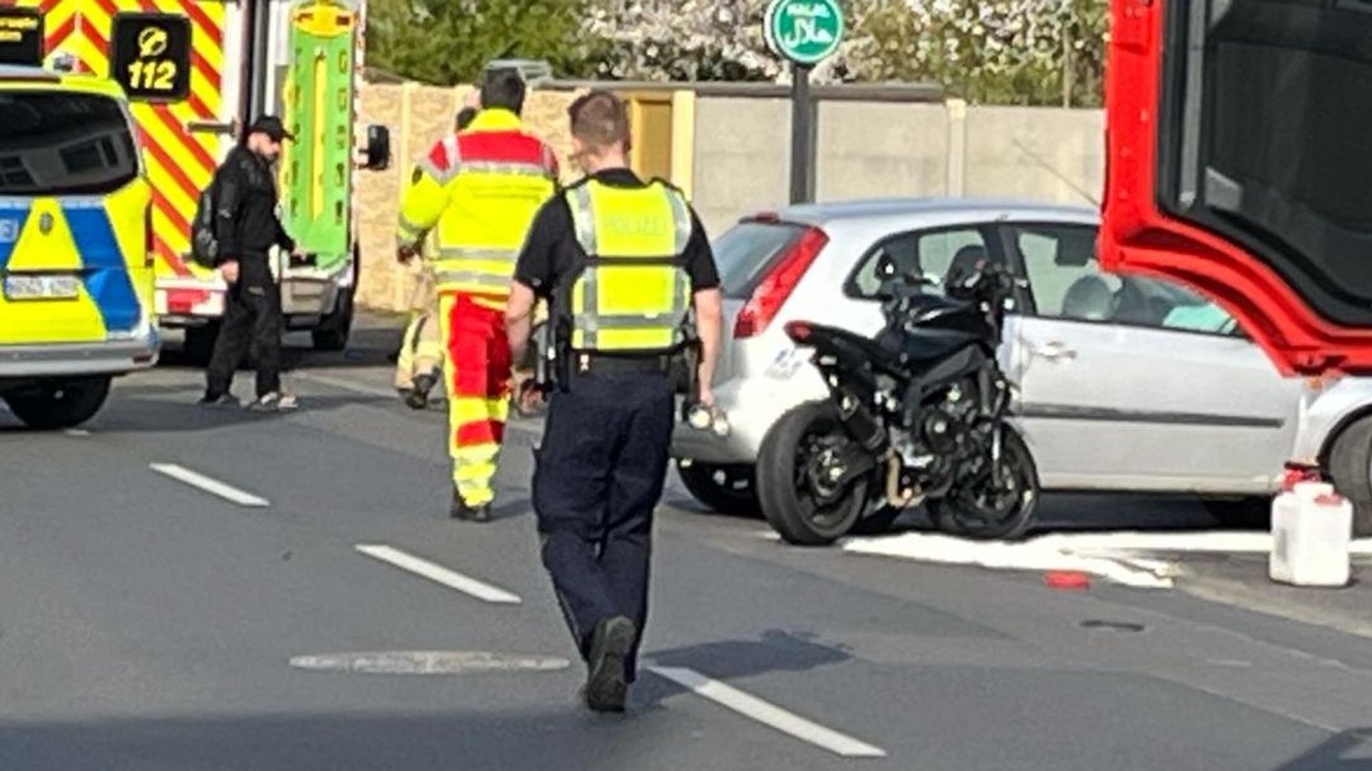 Das Bild zeigt ein Motorrad neben einem Auto und mehrere Einsatzkräfte von Polizei und Feuerwehr.