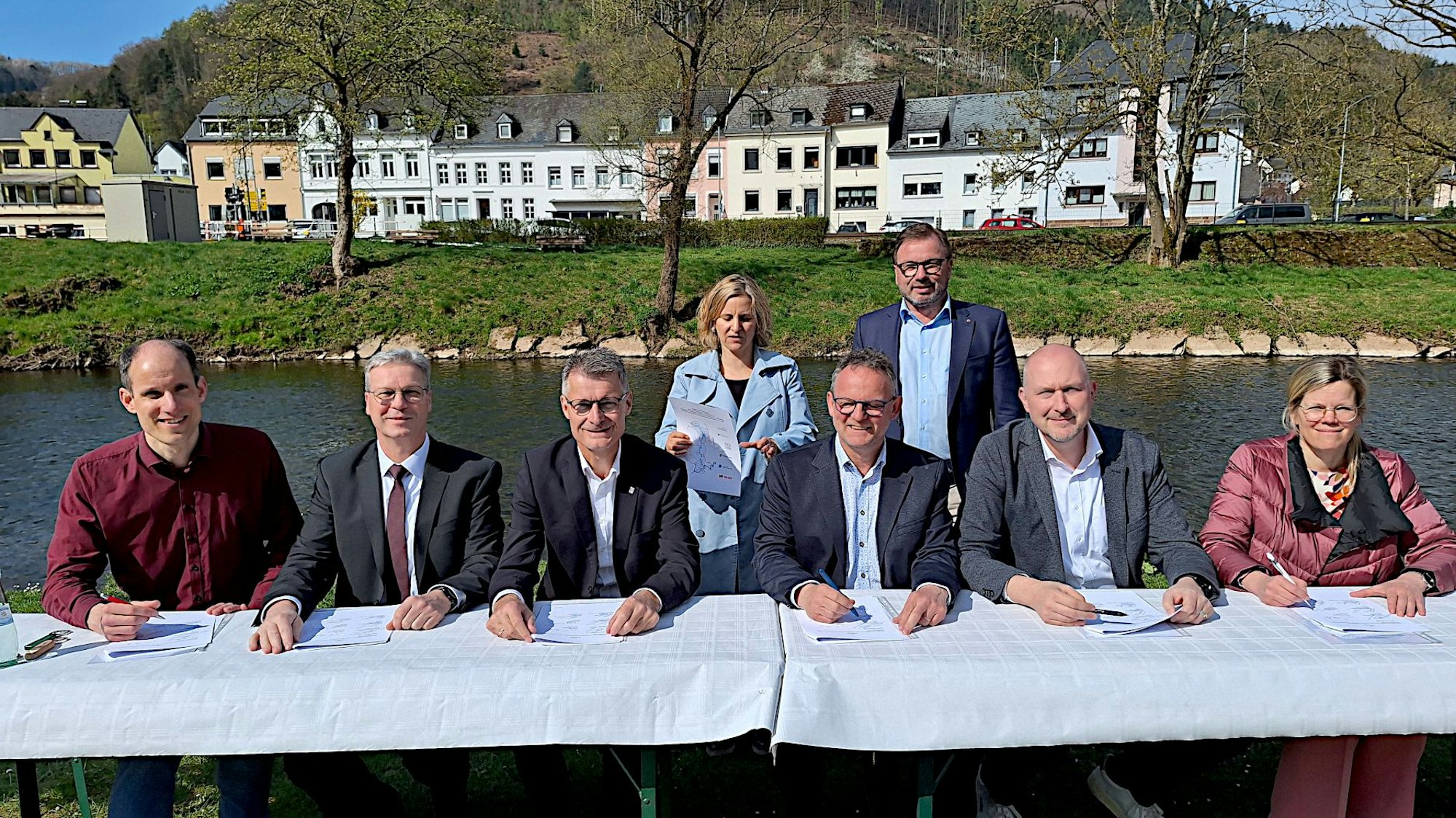 Fünf Männer und eine Frau sitzen an einem Tisch vor einem Fluss. Hinter ihnen stehen ein Mann und eine Frau. Im Hintergrund stehen Häuser und Bäume.