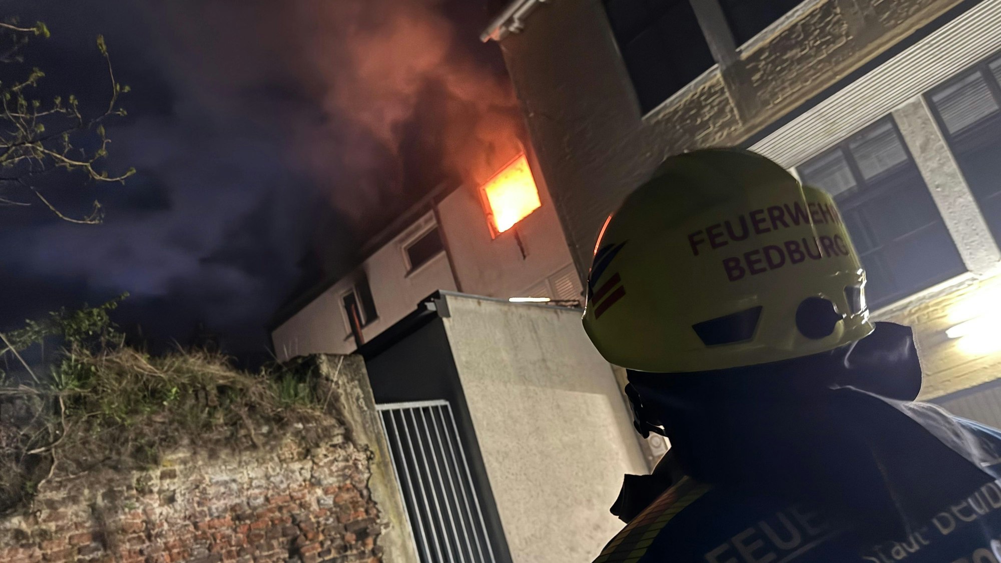 Das Bild zeigt ein Fenster aus der brennenden Wohnung. Davor eines der Einsatzkräfte, auf dem Helm die Aufschrift „Feuerwehr Bedburg“