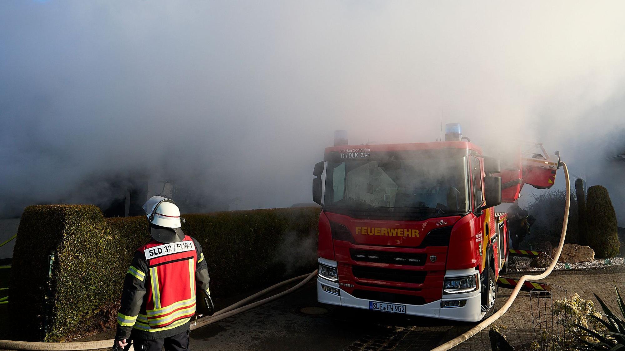 Über einer Hecke und einem Feuerwehrfahrzeug stehen dichte Rauchwolken.