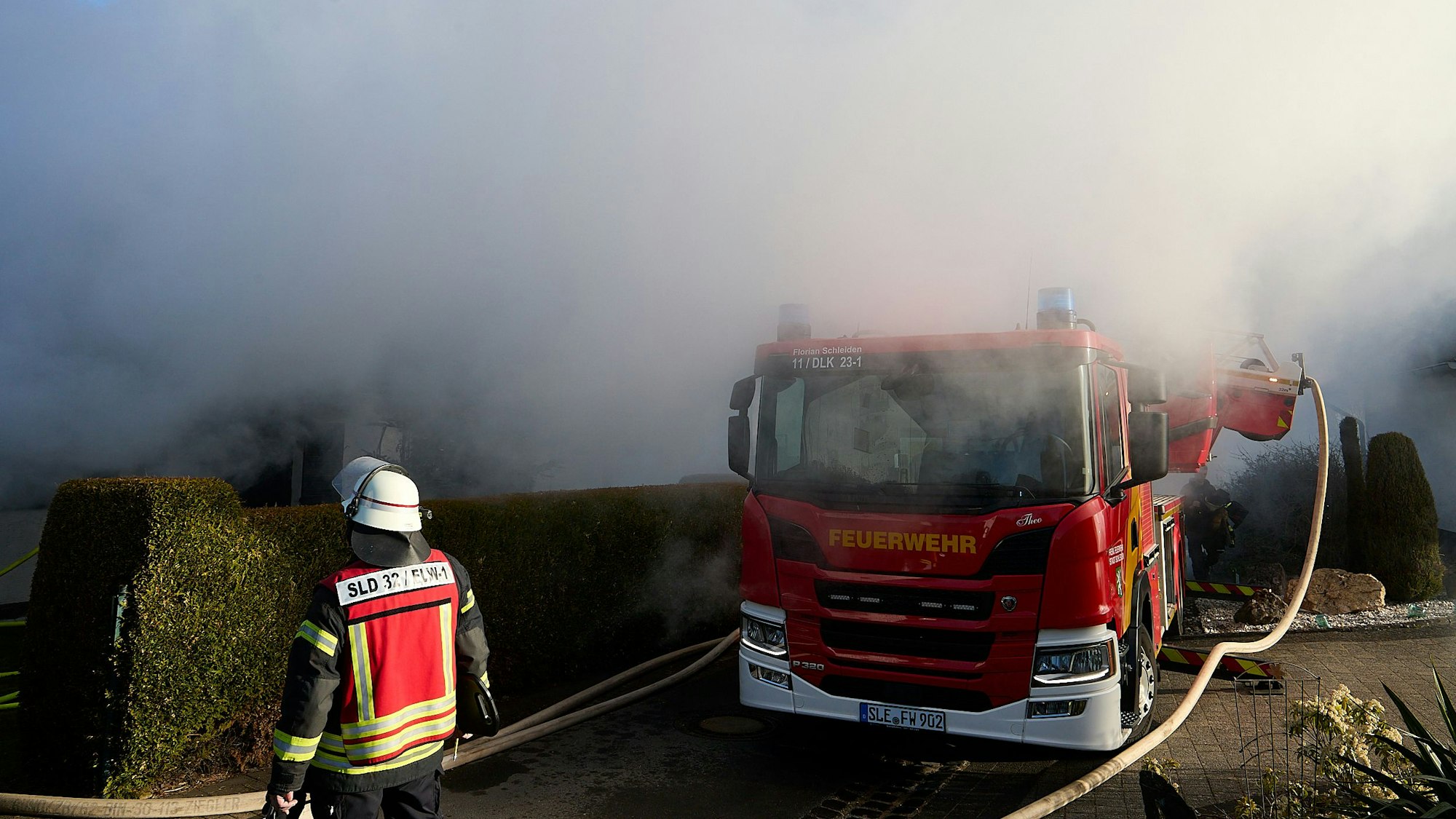 Über einer Hecke und einem Feuerwehrfahrzeug stehen dichte Rauchwolken.