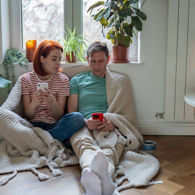 Eine Frau und ein Mann sitzen in ihrer Wohnung. Beide halten ein Smartphone in der Hand, die Frau schaut auf das Smartphone des Mannes.