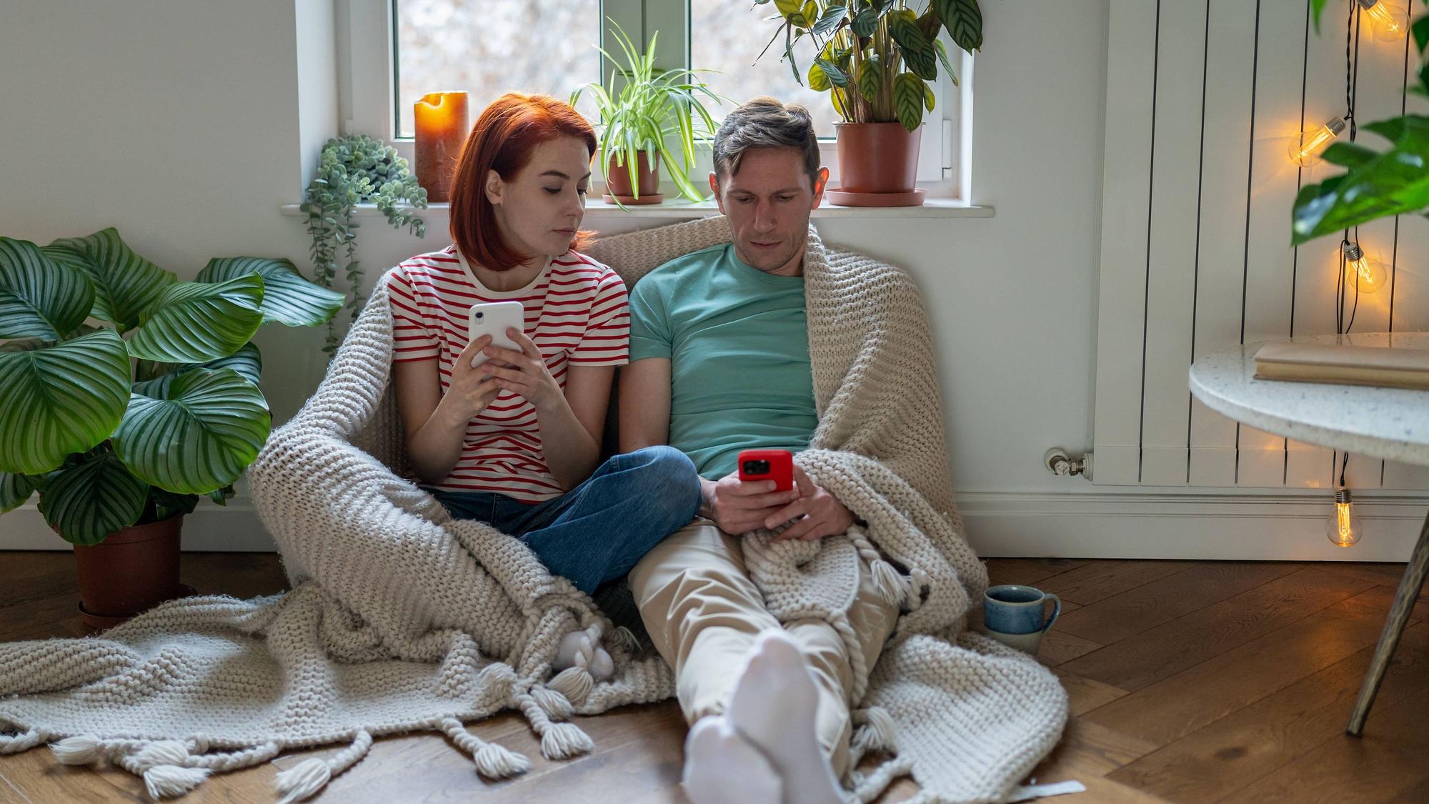 Eine Frau und ein Mann sitzen in ihrer Wohnung. Beide halten ein Smartphone in der Hand, die Frau schaut auf das Smartphone des Mannes.