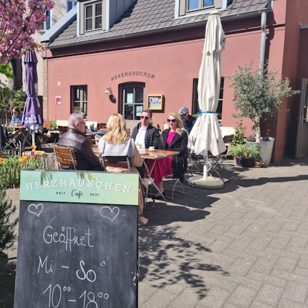 Auf einer Café-Terrasse vor einem Terrakotta-roten Gebäude sitzen viele Menschen an Tischen. Im Vordergrund steht eine Tafel, auf der steht: Geöffnet Mi.–So., 10–18 Uhr.
