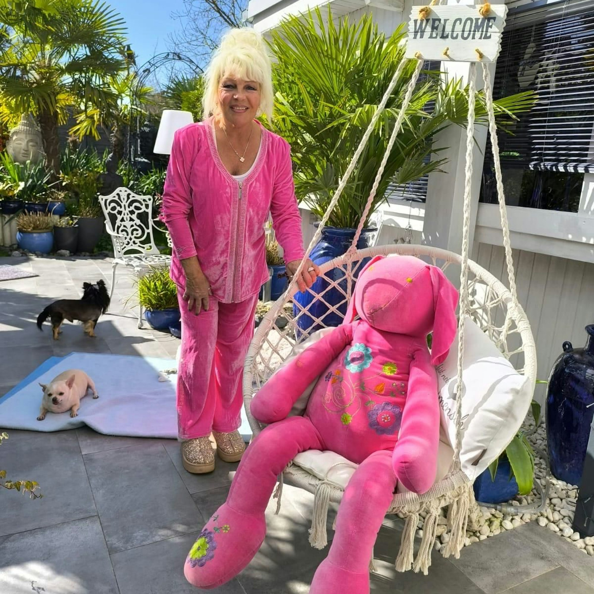 Heike Kleindienst, in rosa gekleidet, steht in ihrem Garten. Neben ihr eine Sitzschaukel mit einem riesigen rosa Plüschhasen.