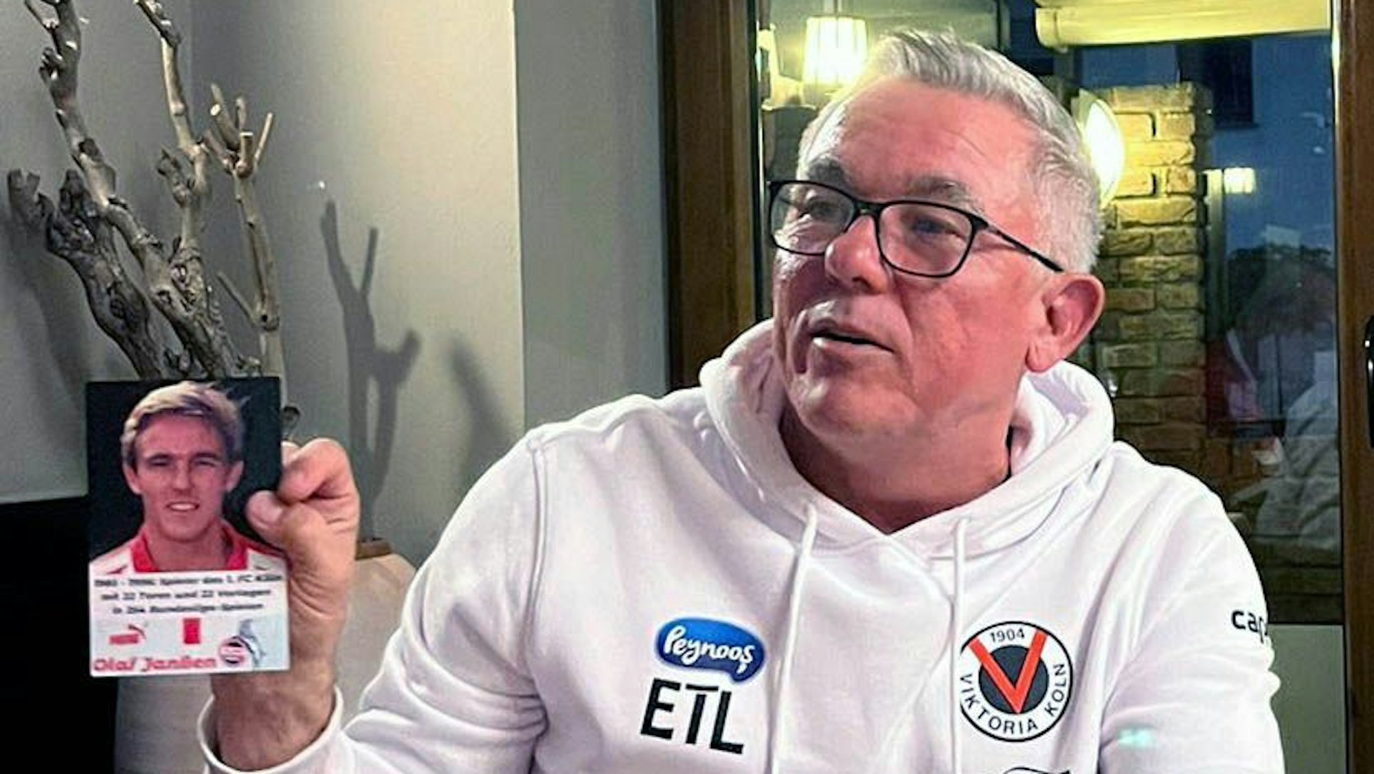 Olaf Janßen trägt einen weißen Pullover mit dem Logo von Viktoria Köln. In der Hand hält er eine Autogrammkarte aus seiner Zeit als Spieler beim 1. FC Köln.