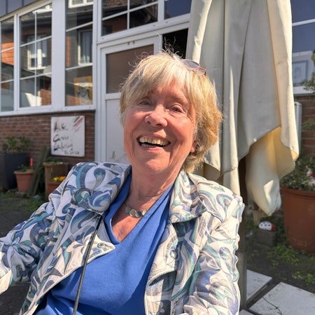 Eine ältere Dame, die vor einem Gebäude auf einer Terrasse sitzt, lacht herzhaft in die Kamera.