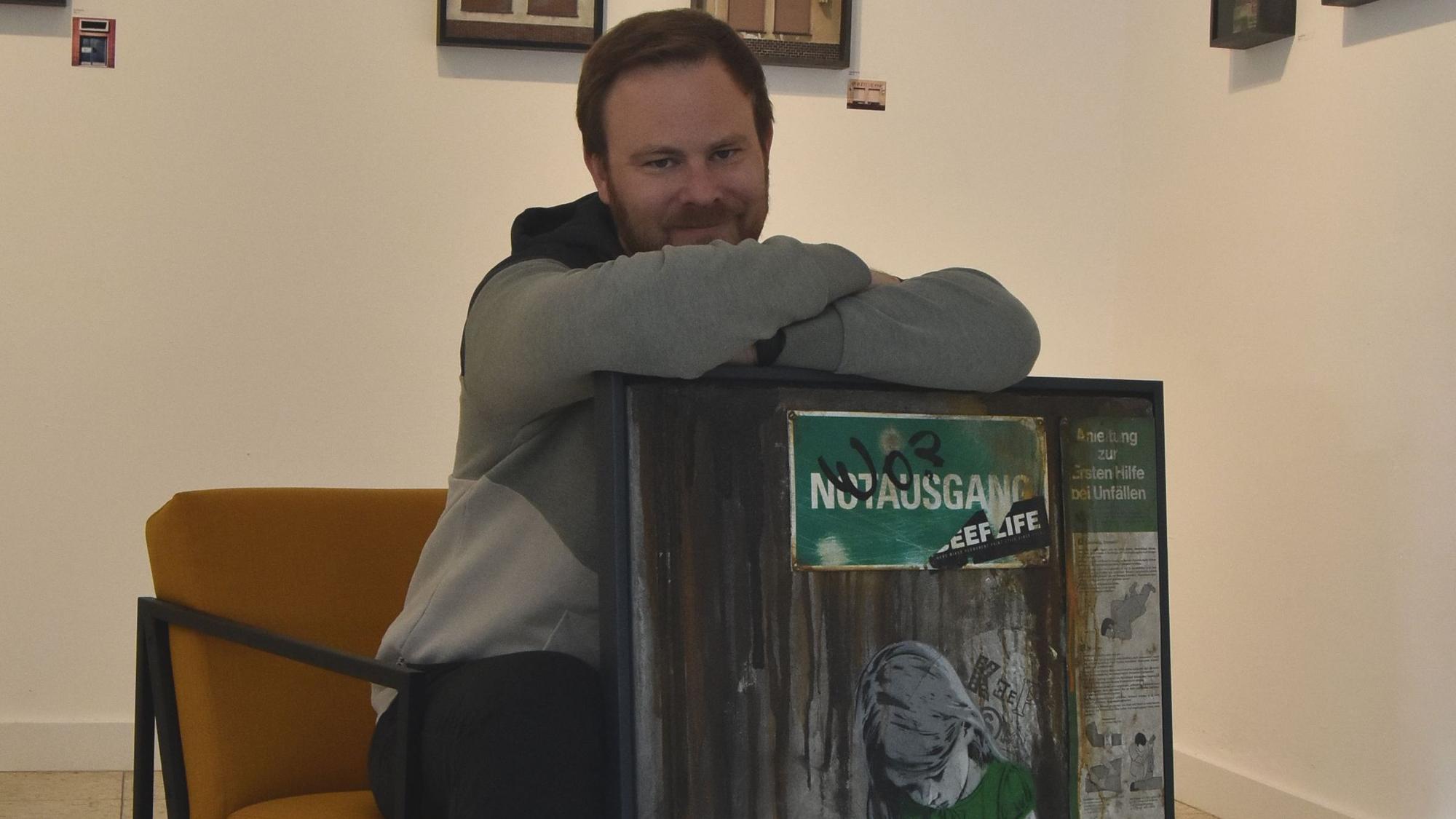 Ein Mann sitzt auf einem senfgelben Sessel und lehnt sich auf ein Graffiti-Werk.