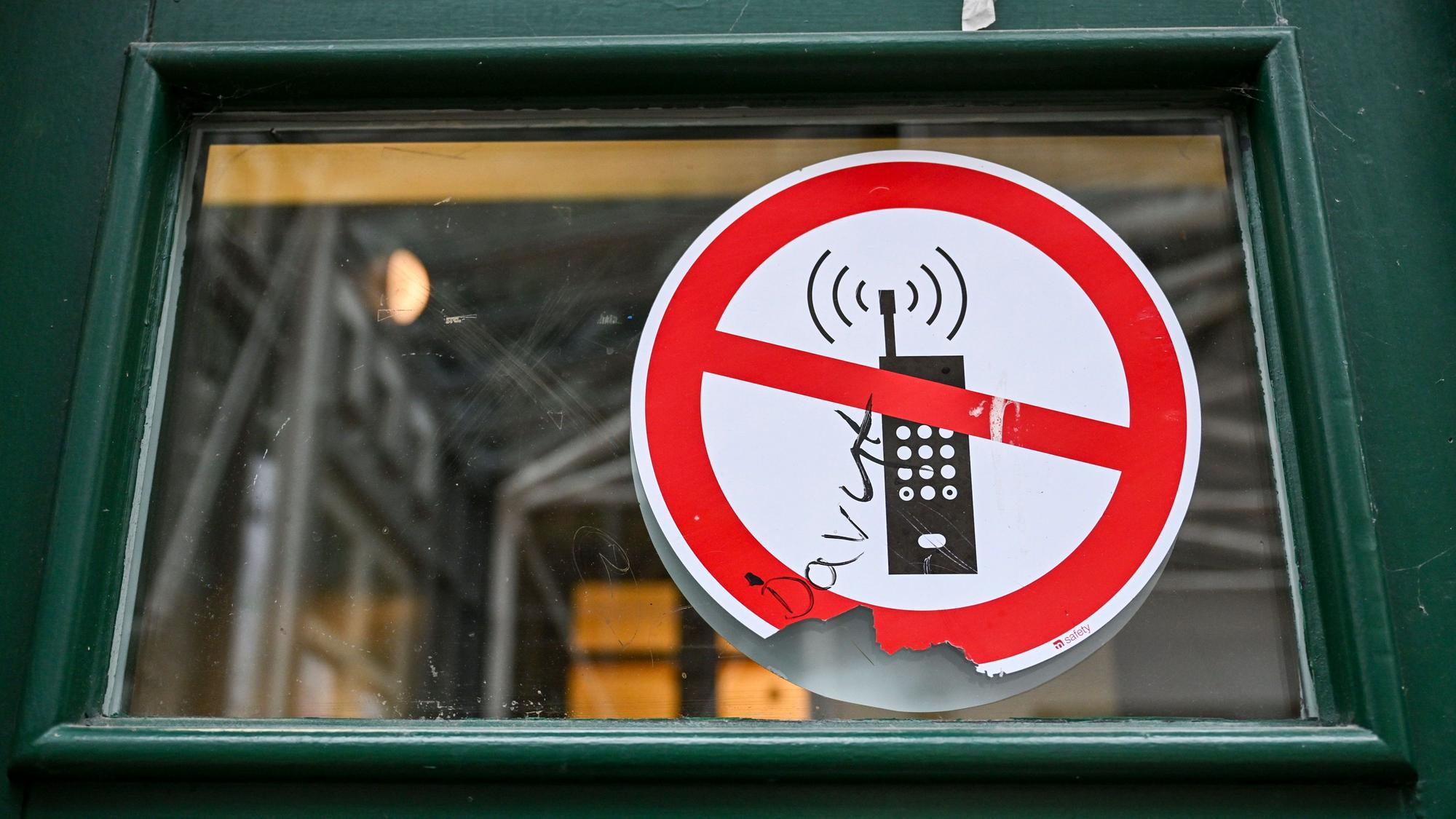 Ein Schild mit einem durchgestrichenen Handy-Symbol weist in einer Schule auf das Handyverbot hin. B