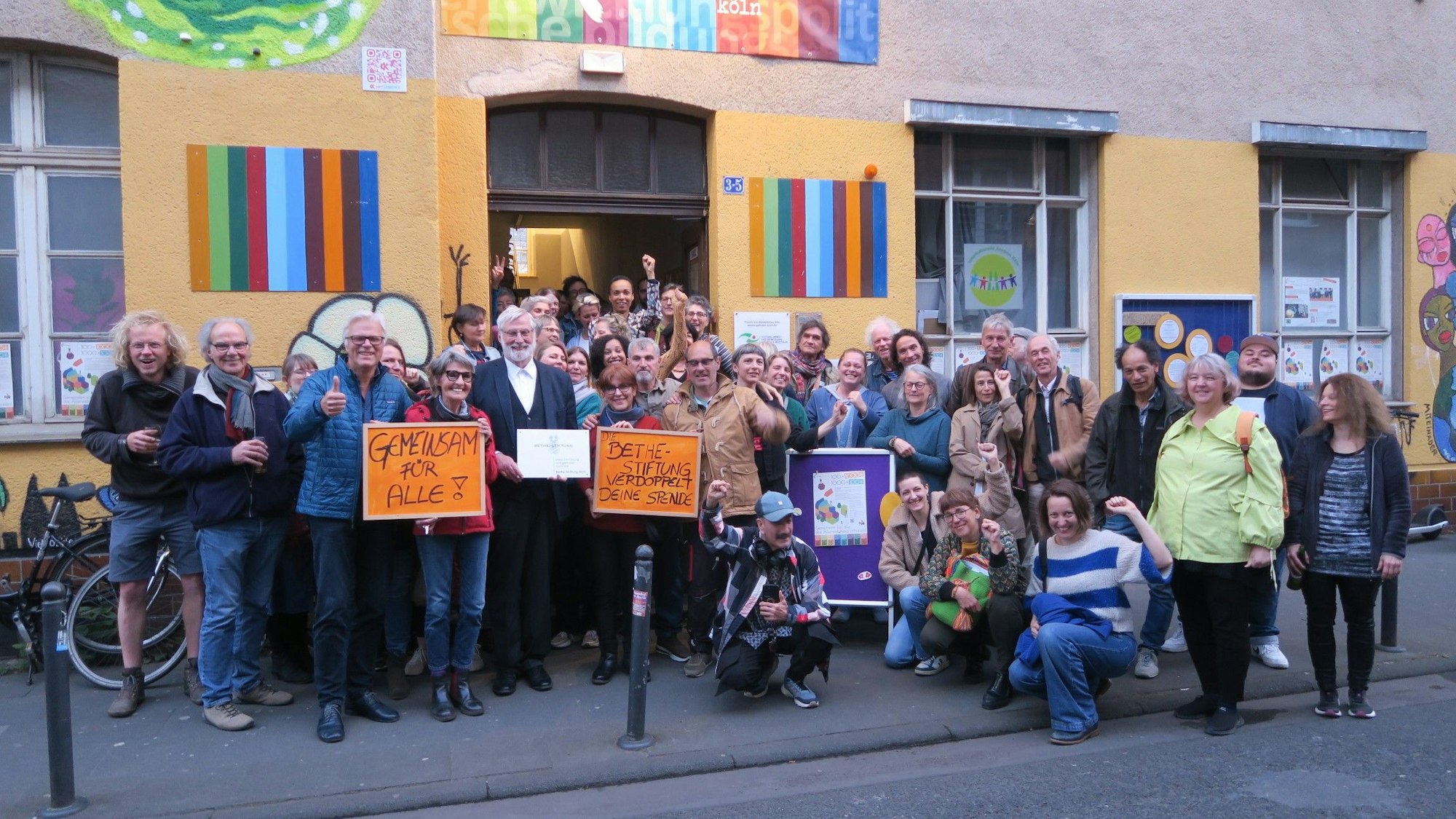 Eine große Menschengruppe steht vor einem gelb verputzten Gebäude mit bunten Plakaten links und rechts von der Tür.