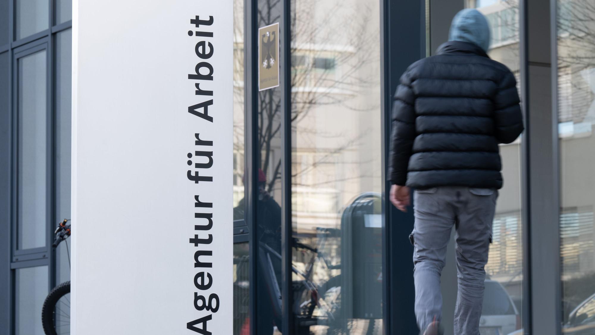 Ein Mann geht hinter einer Stele mit der Aufschrift „Agentur für Arbeit“.